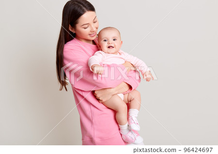 Dark haired young mother hugging her daughter, baby smiling, mommy looking at infant, wearing casual pink sweater and white pants, isolated over light background. Dark haired young mother hugging her daughter, baby smiling, mommy looking at infant, wearing casual pink sweater and white pants, isolated over light background. 96424697