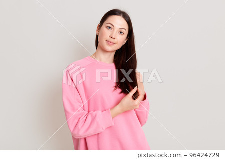 Portrait of charming dark haired female wearing pink sweatshirt, looking at camera with calm expression and cute smile, touching her hair with hands, isolated over white background. Portrait of charming dark haired female wearing pink sweatshirt, looking at camera with calm expression and cute smile, touching her hair with hands, isolated over white background. 96424729