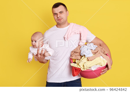Single father overwhelmed by taking care of everything alone, take care of new born baby and doing laundry, wearing casually, stands isolated over yellow background, holds basin with clean clothing. Single father overwhelmed by taking care of everything alone, take care of new born baby and doing laundry, wearing casually, stands isolated over yellow background, holds basin with clean clothing. 96424842