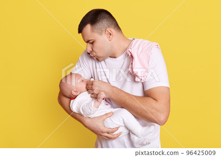 Young Caucasian father gives to baby soother, looking at her daughter or son with great love, guy wearing white t shirt, posing isolated over yellow background. 96425009