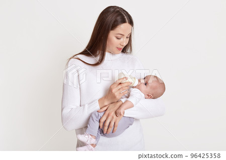 Mother feeding her baby from feeding bottle, dark haired female wearing sweater hold baby in hands and looking at her kid, posing isolated over white background. Mother feeding her baby from feeding bottle, dark haired female wearing sweater hold baby in hands and looking at her kid, posing isolated over white background. 96425358