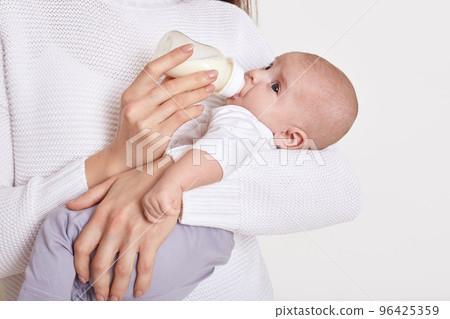 Mother feeding from bottle her baby isolated over white background, little infant in mommy hands, eating milk, kid in white shirt and grey pants. Childhood and motherhood. Mother feeding from bottle her baby isolated over white background, little infant in mommy hands, eating milk, kid in white shirt and grey pants. Childhood and motherhood. 96425359
