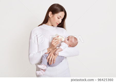 Mother feeding from bottle her baby isolated over white background, lady with brown hair wearing sweater holding infant and looking on her, young mommy with child. Mother feeding from bottle her baby isolated over white background, lady with brown hair wearing sweater holding infant and looking on her, young mommy with child. 96425360