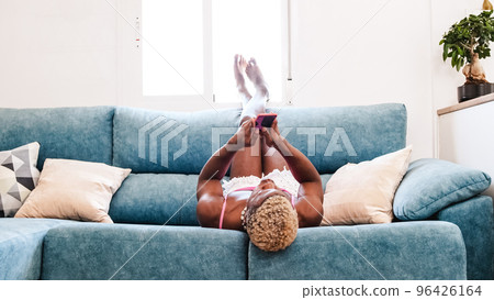 Transgender person using a mobile phone while lying upside down on a sofa at home. 96426164