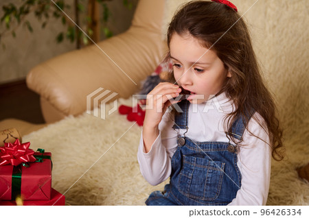 Caucasian cute child, adorable lovely baby girl with beautiful long hair, wearing white shirt and blue denim overalls, eating chocolates while sitting on sofa at home with Christmas gifts on a table Caucasian cute child, adorable lovely baby girl with beautiful long hair, wearing white shirt and blue denim overalls, eating chocolates while sitting on sofa at home with Christmas gifts on a table 96426334