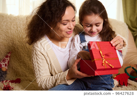 Multi-ethnic pretty woman in warm beige sweater, a loving mother hugging her adorable lovely daughter, while giving her a cute Christmas present. Happy childhood. Winter and New Year's holidays Multi-ethnic pretty woman in warm beige sweater, a loving mother hugging her adorable lovely daughter, while giving her a cute Christmas present. Happy childhood. Winter and New Year's holidays 96427628