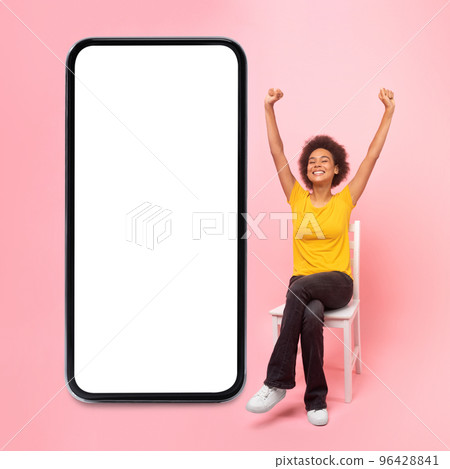 Glad millennial african american curly woman rejoices, makes gesture of win, success, sits on chair, raises hands up Glad millennial african american curly woman rejoices, makes gesture of win, success, sits on chair, raises hands up 96428841