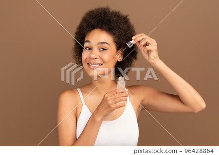 Glad millennial black curly female in white top apply anti-aging collagen serum, isolated on brown background Glad millennial black curly female in white top apply anti-aging collagen serum, isolated on brown background 96428864