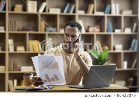 Irritated arab businessman working from home, sitting at desk with laptop, holding documents and having phone call Irritated arab businessman working from home, sitting at desk with laptop, holding documents and having phone call 96428879