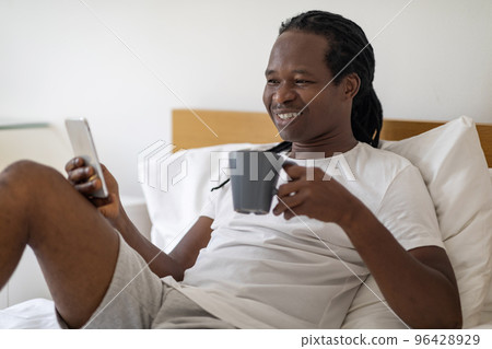 Portrait Of Smiling Black Man Resting In Bed With Smartphone And Coffee 96428929
