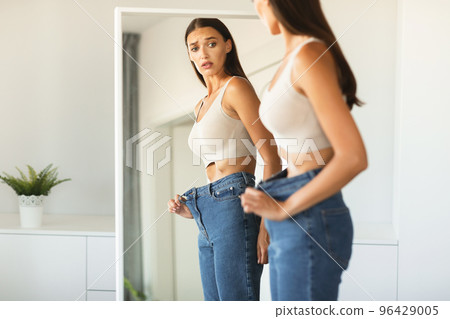 Shocked young woman wearing too big jeans after successful weight loss, posing and looking at her reflection in mirror Shocked young woman wearing too big jeans after successful weight loss, posing and looking at her reflection in mirror 96429005