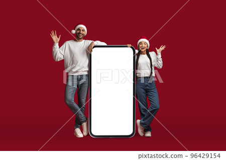Excited Middle Eastern Couple Wearing Santa Hats Posing Near Big Blank Smartphone 96429154