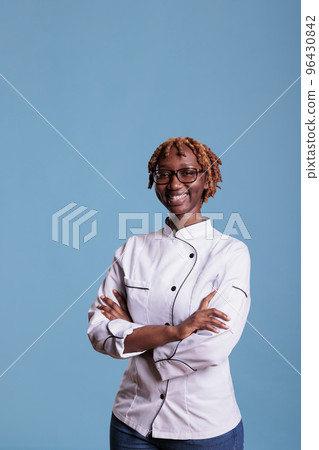 African american professional female chef posing in white uniform in studio shot. Happy cook with curly hair looking at camera with arms crossed against blue background. African american professional female chef posing in white uniform in studio shot. Happy cook with curly hair looking at camera with arms crossed against blue background. 96430842