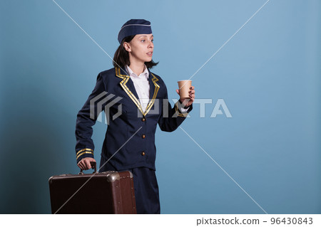 Flight attendant in professional aviation uniform running late, carrying suitcase. Stewardess with baggage walking in airport, woman holding cup of coffee, side view medium shot Flight attendant in professional aviation uniform running late, carrying suitcase. Stewardess with baggage walking in airport, woman holding cup of coffee, side view medium shot 96430843