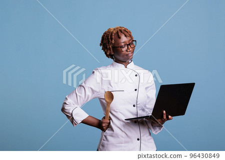 Food industry worker in kitchen uniform using computer to inspire a mediterranean meal. Chef with laptop looking for recipes of dishes on internet while standing in studio. Food industry worker in kitchen uniform using computer to inspire a mediterranean meal. Chef with laptop looking for recipes of dishes on internet while standing in studio. 96430849