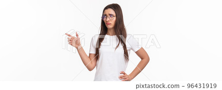 Waist-up portrait of uneasy, sad and gloomy young brunette woman in glasses, looking and pointing finger right with sulking unhappy face, express jealousy or regret, grieving white background 96431919