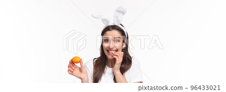 Easter, holidays and spring concept. Close-up portrait of silly and cute, happy giggling girl in rabbit ears, made her first colored egg on Easter day, biting finger, looking camera joyful Easter, holidays and spring concept. Close-up portrait of silly and cute, happy giggling girl in rabbit ears, made her first colored egg on Easter day, biting finger, looking camera joyful 96433021