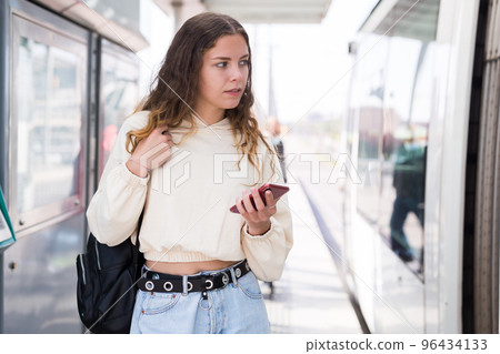 Portrait of a girl standing at a stop next to a tram, holding a mobile phone Portrait of a girl standing at a stop next to a tram, holding a mobile phone 96434133