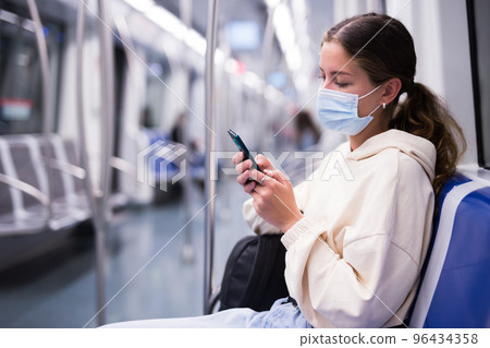 Active girl in a protective mask riding on a subway train with a mobile phone Active girl in a protective mask riding on a subway train with a mobile phone 96434358