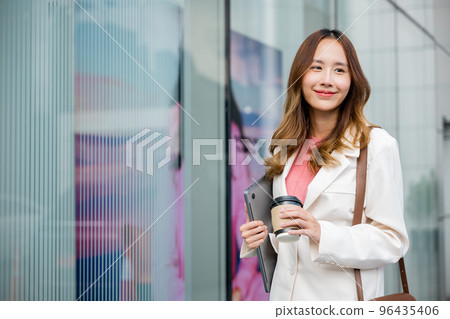Young business woman wearing white suit jacket and smile while go to work outdoor mirror building background. Confident Businesswoman with cup of coffee holding Laptop walking outside office building Young business woman wearing white suit jacket and smile while go to work outdoor mirror building background. Confident Businesswoman with cup of coffee holding Laptop walking outside office building 96435406