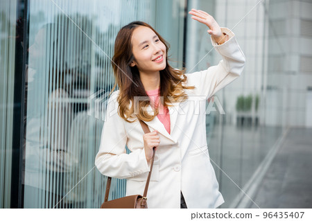 Portrait of confidence young businesswoman standing outside office building in city. Happy woman wearing white suit jacket with brown bag at sunlight outdoors. Portrait of confidence young businesswoman standing outside office building in city. Happy woman wearing white suit jacket with brown bag at sunlight outdoors. 96435407