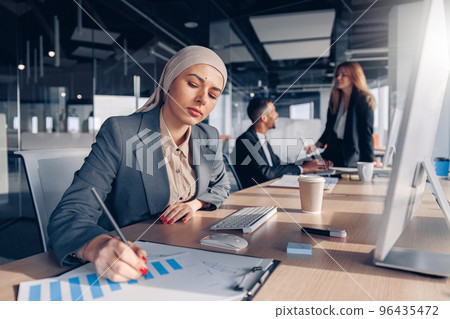 Focused muslim businesswoman in hijab working with documents while sitting in modern office Focused muslim businesswoman in hijab working with documents while sitting in modern office 96435472