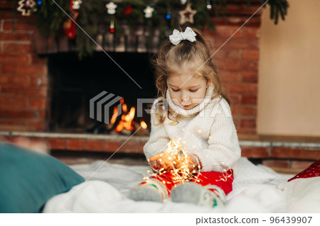 Cute little girl sitting by the fireplace with a Christmas garland.Christmas portrait, cozy style. The concept of Christmas and New Year 96439907