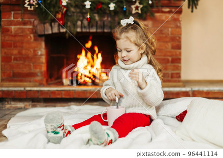 A baby girl drinks hot chocolate by the fireplace at home. A little girl on Christmas Eve or New Year's Eve at home. The concept of Christmas and New Year A baby girl drinks hot chocolate by the fireplace at home. A little girl on Christmas Eve or New Year's Eve at home. The concept of Christmas and New Year 96441114