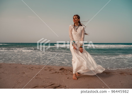 Model in boho style in a white long dress and silver jewelry on the beach. Her hair is braided, and there are many bracelets on her arms. 96445982