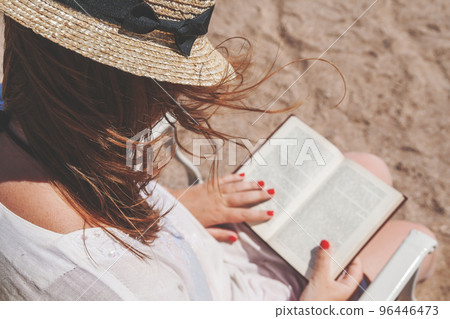 Young adult woman with a hat on the beach reading a book Young adult woman with a hat on the beach reading a book 96446473