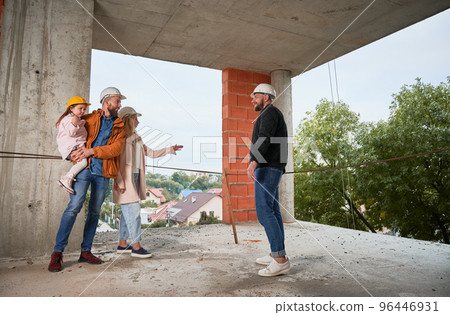 Man and woman with child standing inside apartment building under construction and talking with builder. Family future homeowners discussing building process with construction worker. 96446931