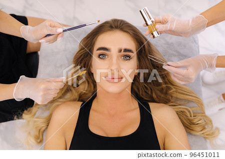 Top view of four hand of beauticians making eyelash and brow lamination procedures for young blond hair model. Professional beauty studio Top view of four hand of beauticians making eyelash and brow lamination procedures for young blond hair model. Professional beauty studio 96451011