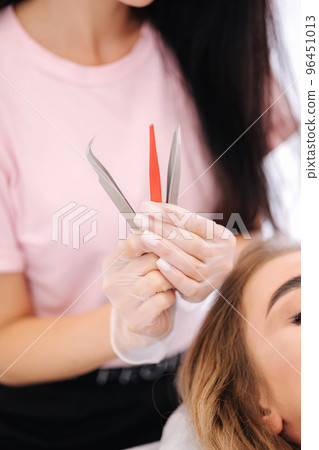 Woman doing eyebrow correction and lamination in beauty studio. Blond hair model has staining, curling, laminating and extension for brow. Beauticiaon hold tweezers 96451013