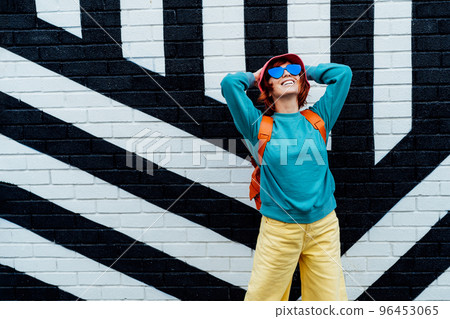 Happy emotional hipster fashion young woman in bright clothes, heart shape sun glasses and bucket hat posing on the painted brick wall background. Urban city street fashion. Selective focus. 96453065