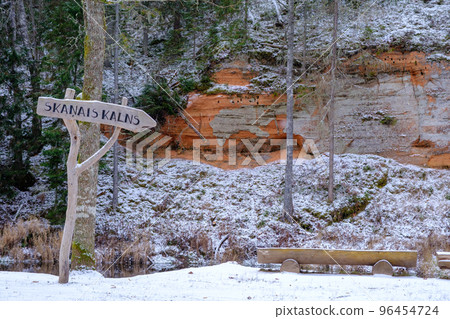 A view of the red sandstone Skanaiskalns cliffs near Salacas. Signpost and wooden bench by the river. Skanaiskalna Nature Park in November in Mazsalac, Latvia. A view of the red sandstone Skanaiskalns cliffs near Salacas. Signpost and wooden bench by the river. Skanaiskalna Nature Park in November in Mazsalac, Latvia. 96454724