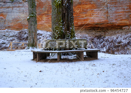 A bench in the shape of a snowy circle in a nature park. Encircles the trunk of an oak tree. In the background, a large red sandstone rock. Skanaiskalna National Park, Latvia. A bench in the shape of a snowy circle in a nature park. Encircles the trunk of an oak tree. In the background, a large red sandstone rock. Skanaiskalna National Park, Latvia. 96454727