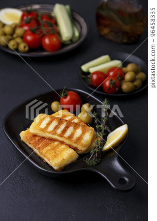 Grilled halloumi cheese in a black pan with olives, tomatoes, cucumbers, thyme and lemon. Traditional cheese of the Eastern Mediterranean. Black background, selective focus, close-up. 96458335