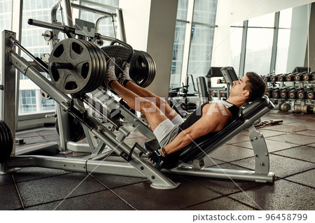 Young man is engaged on a modern simulator in the gym. Male athlete is doing exercises for legs with training machine. Young man is engaged on a modern simulator in the gym. Male athlete is doing exercises for legs with training machine. 96458799