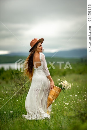 A middle-aged woman in a white dress and brown hat stands on a green field and holds a basket in her hands with a large bouquet of daisies. In the background there are mountains and a lake. 96459126