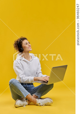 Side view of pretty girl with curly hair working at laptop. Blonde young female listening to music in earphones, looking up, smiling, Isolated on yellow studio background. 96460247