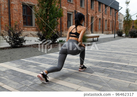 Fitness woman stretches her legs doing lunges on the street near a brick building 96462317