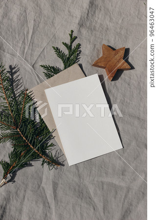 Natural Christmas stationery still life. Blank greeting card, invitation mock up with wooden star ornament and fir, cypress tree branch in sunlight. Craft envelope, linen tablecloth. Vertical top view Natural Christmas stationery still life. Blank greeting card, invitation mock up with wooden star ornament and fir, cypress tree branch in sunlight. Craft envelope, linen tablecloth. Vertical top view 96463047