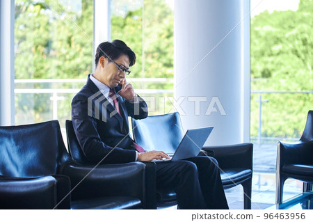 A businessman in a suit making a phone call on a smartphone while using a computer in an airport lobby A businessman in a suit making a phone call on a smartphone while using a computer in an airport lobby 96463956