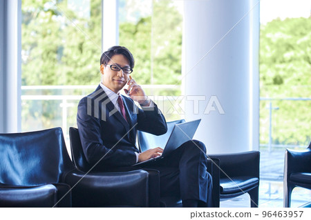 A businessman in a suit making a phone call on a smartphone while using a computer in an airport lobby 96463957