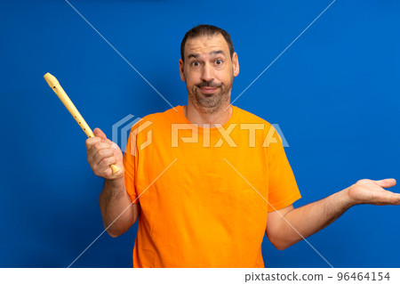 Bearded latino man in orange t-shirt frustrated and pissed off for not being able to play the flute decently, isolated over blue studio background. 96464154