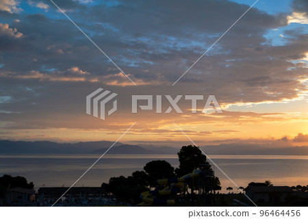 Sunrise From Corfu Island Overlooking Mountains Of Balkan Peninsula Of Greece, Moraitika, Corfu, Greece. In Foreground Are Silhouettes Of Trees, Beach Umbrellas And Hotel Pool. 96464456