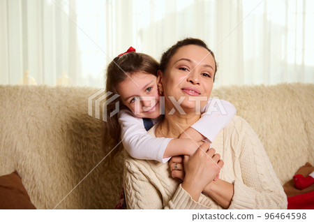 Lovely little girl gently hugging her mother from the back, sitting together on sofa at home and cutely smiling looking at camera. Happy Childhood and Motherhood. Love. Care. Family relationships 96464598