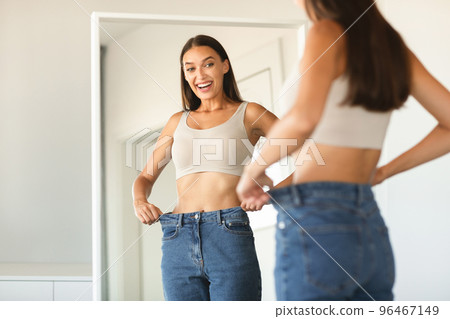 Successful slimming concept. Excited caucasian lady wearing jeans after weight loss, comparing size, posing near mirror Successful slimming concept. Excited caucasian lady wearing jeans after weight loss, comparing size, posing near mirror 96467149