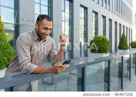 Young handsome Hispanic businessman, African American standing on veranda of office center. Holding phone, playing online games, pointing yes with hand, rejoices at the victory. 96467168