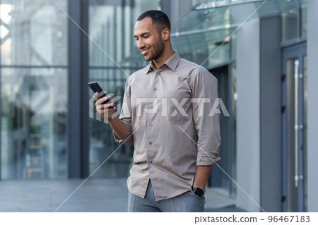 A young handsome Hispanic, African-American man is standing on the street near an office center. He holds the phone in his hand, uses it, dials, smiles. 96467183